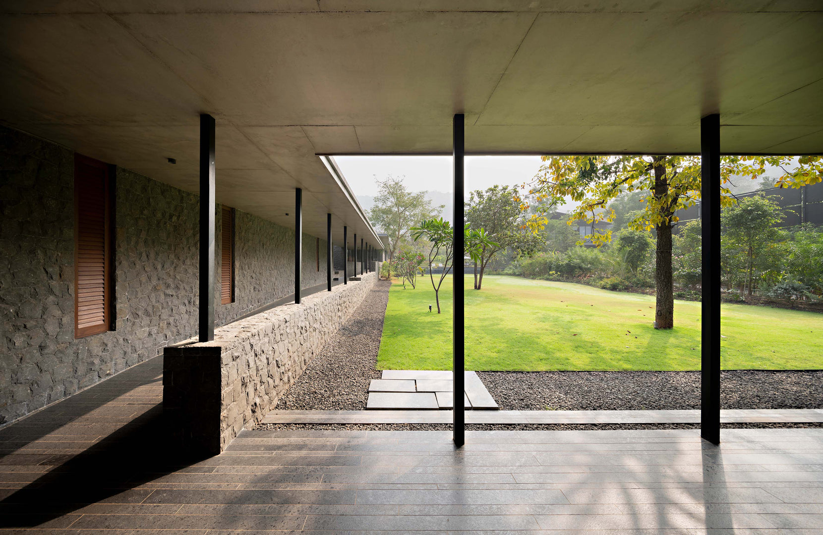 Covered walkway between a rough stone wall and slender steel columns, opening to the lawn