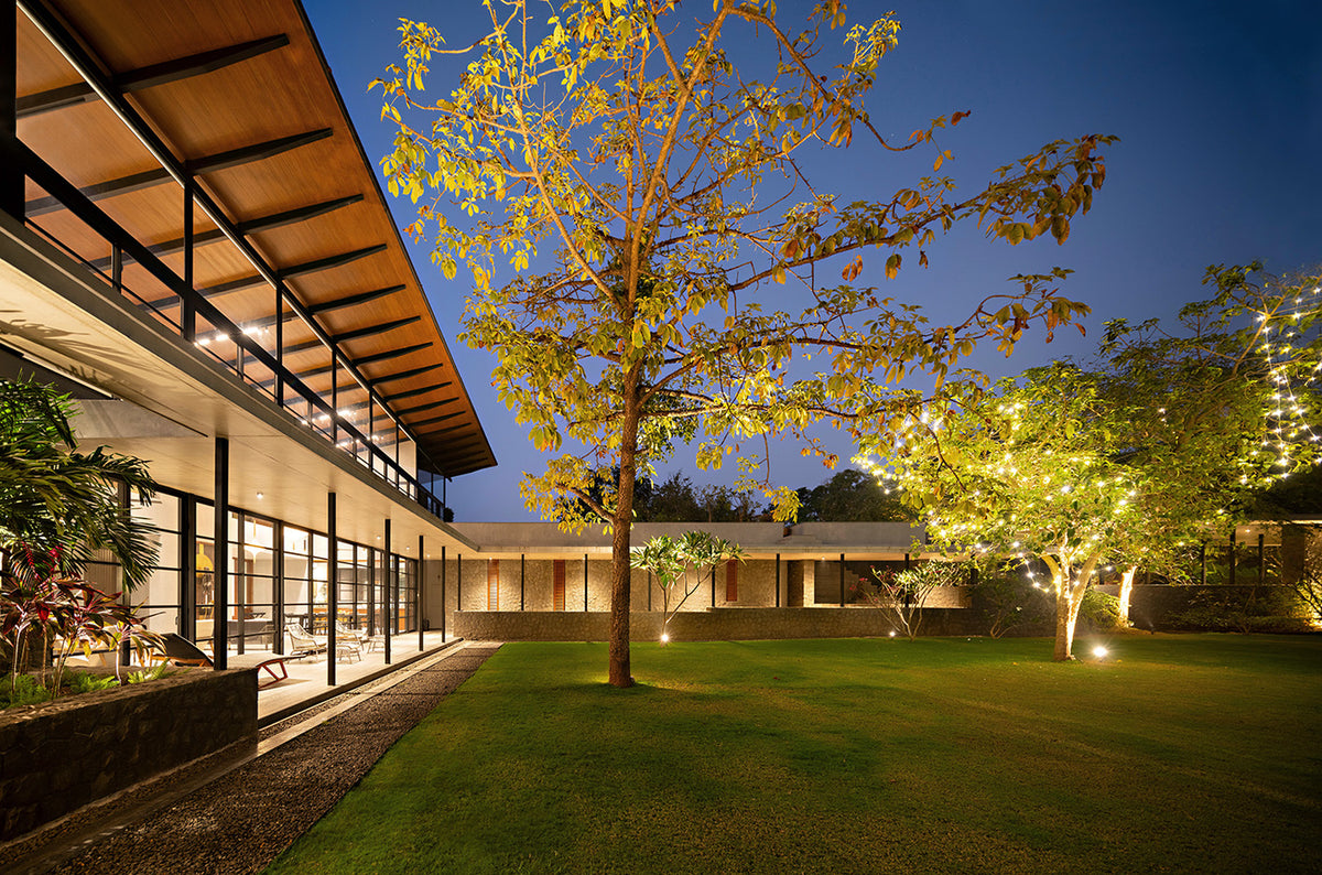 Alibaug Commune at dusk, its lit glazed verandah facing a lawn and a tree strung with lights