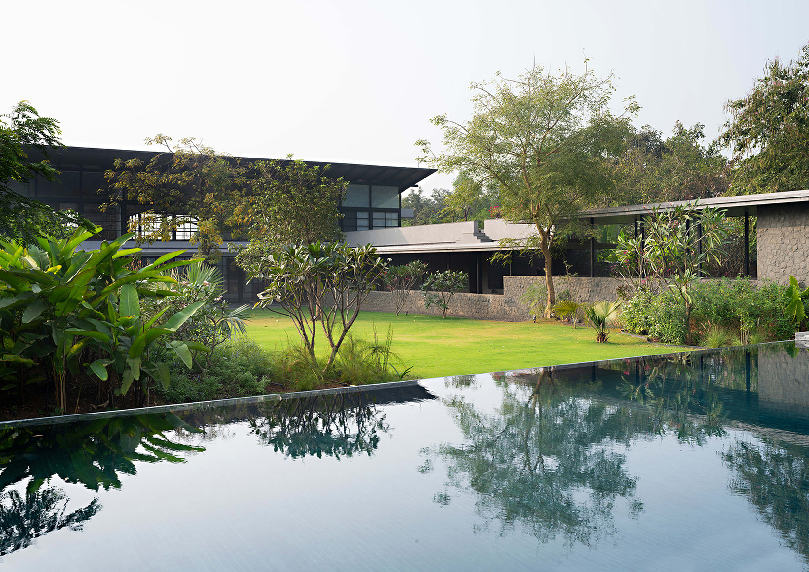 Infinity pool reflecting the lawn and the stone-plinthed pavilion under its steel-framed roof