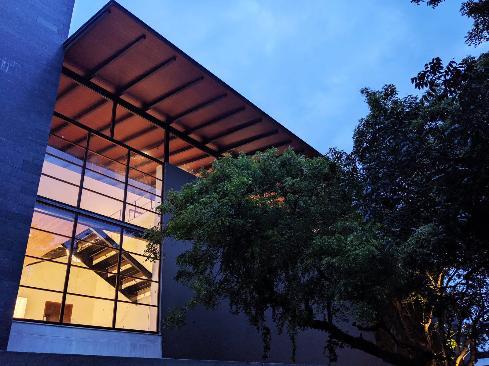 Steel-framed roof overhang from below at dusk, the double-height living space glowing within
