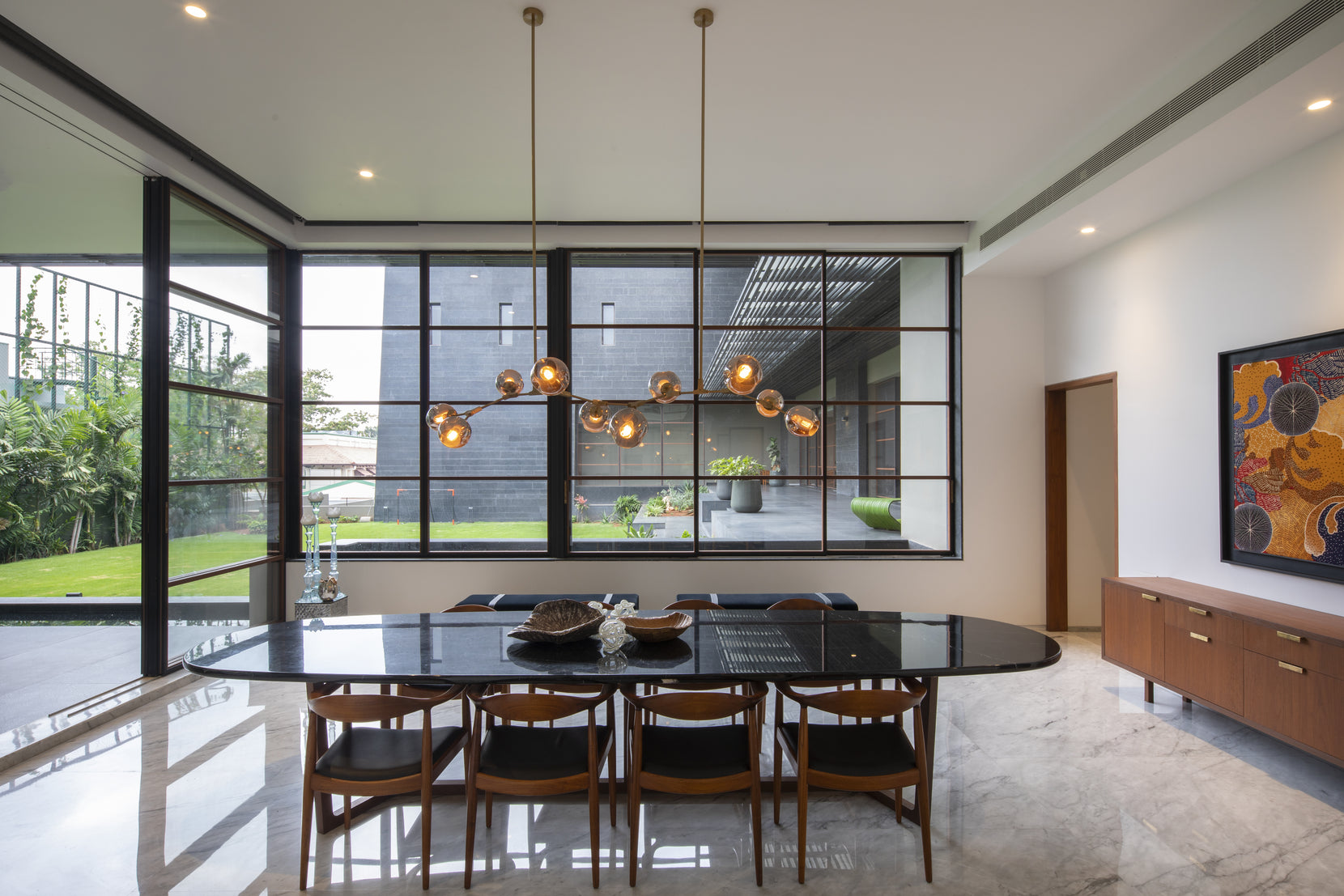 Dining room with an oval black table and brass pendant, garden framed through steel glazing