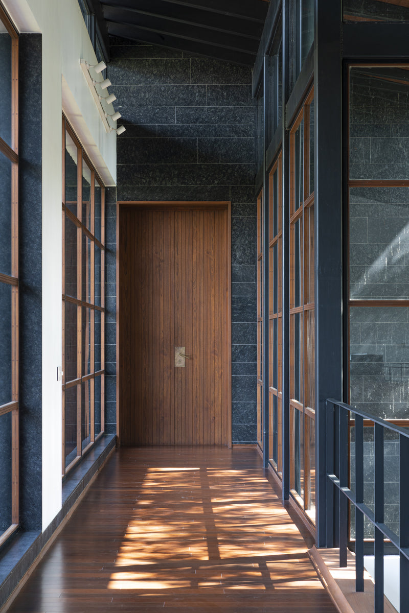 Sunlight patterning the timber floor of a corridor between granite walls, ending at a teak door
