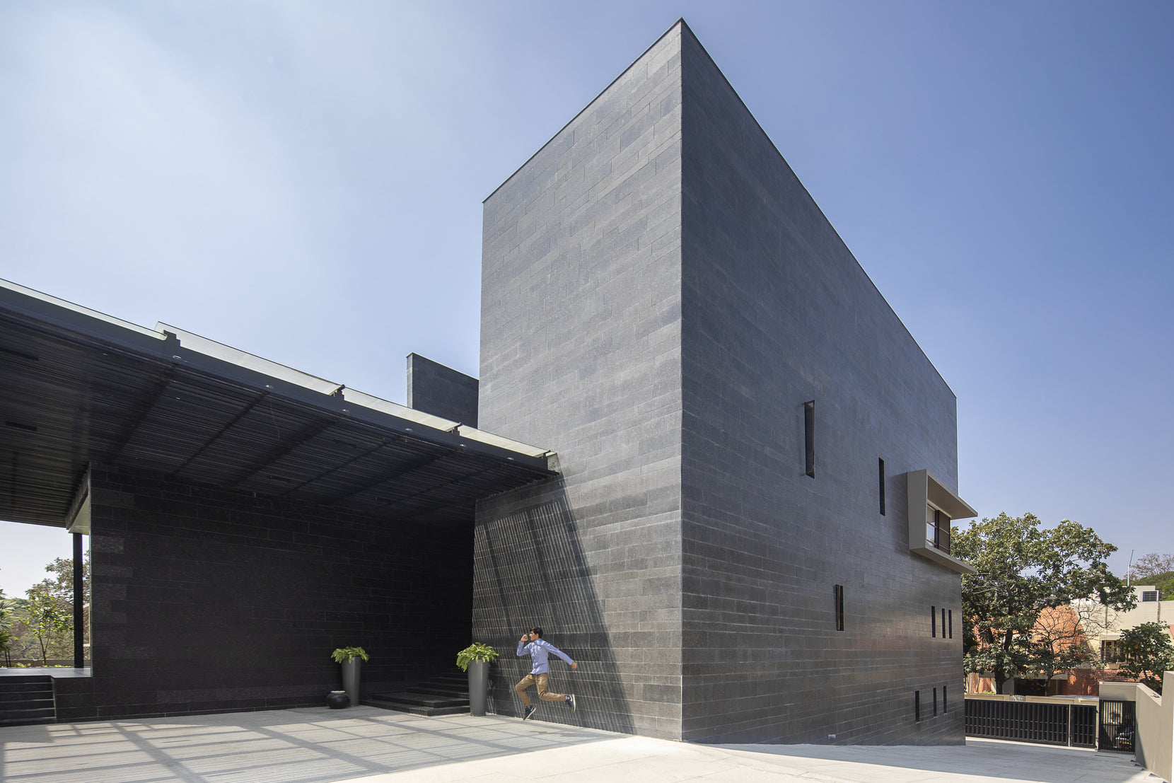 Blank granite volume beside the covered porch, a figure running across the sunlit forecourt