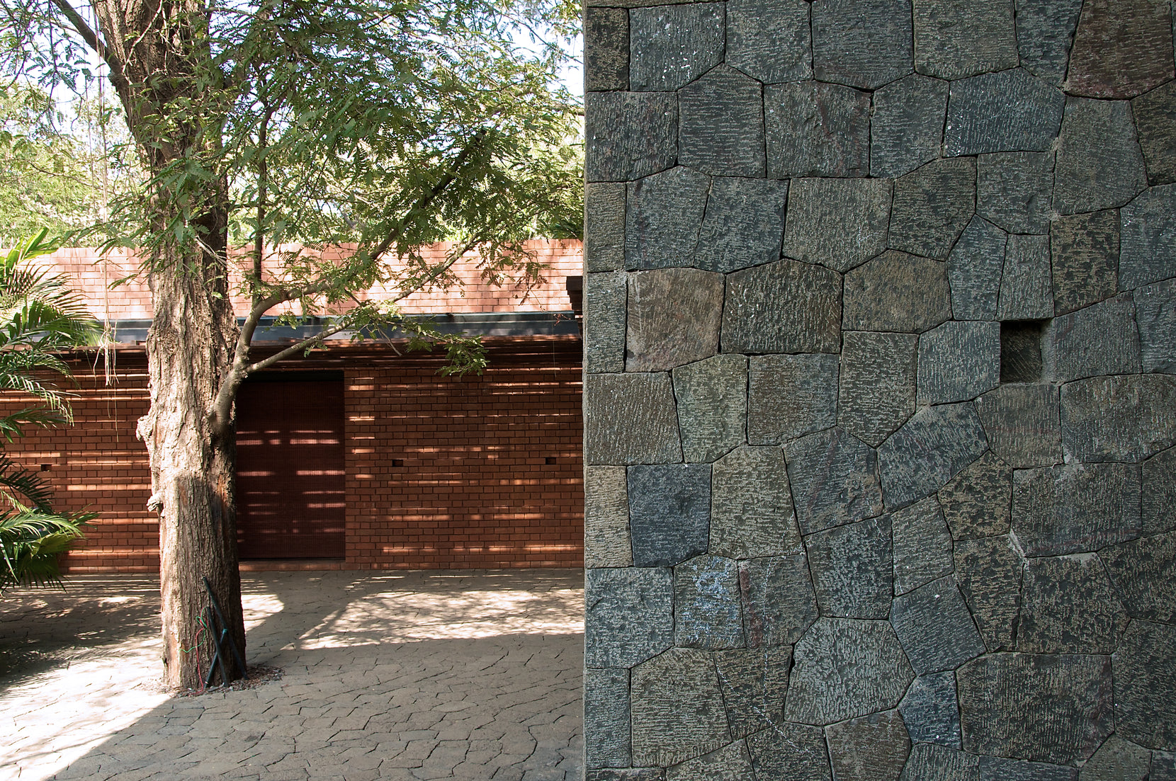 Split composition of a basalt rubble wall and the brick facade behind a mature tree