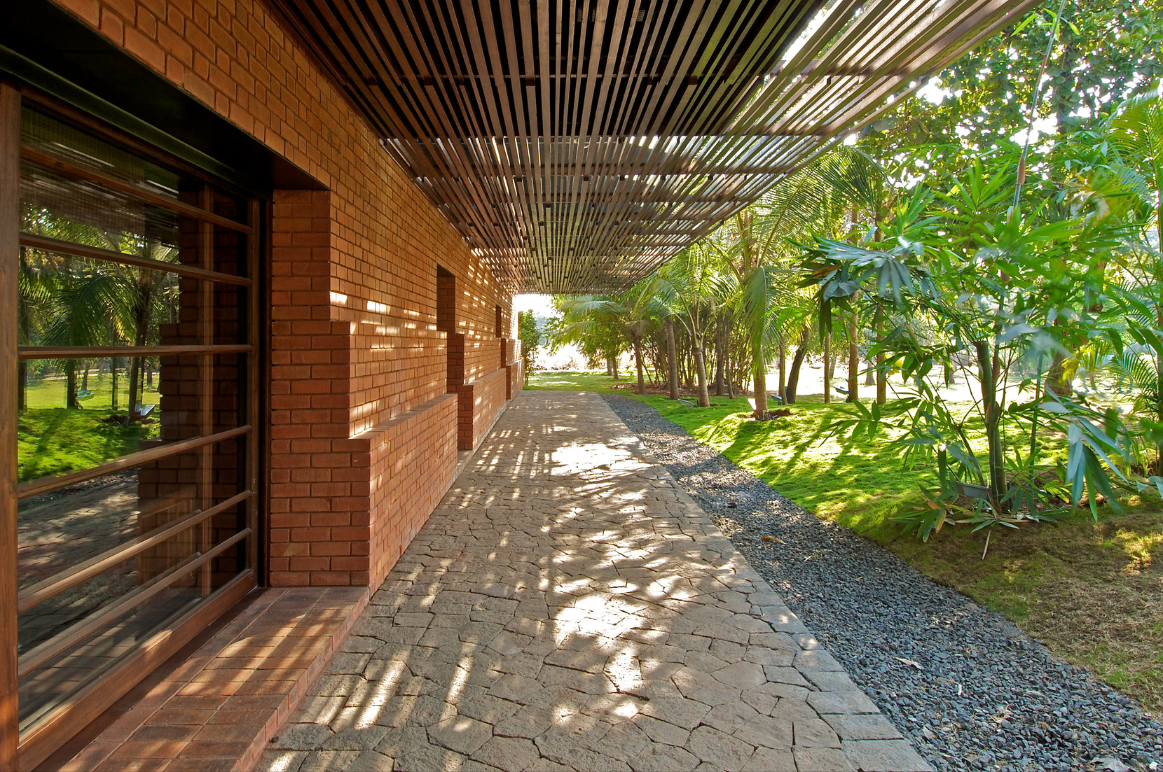 Brick Kiln House, Alibaug: a shaded walkway of stone paving beside brick walls and timber rafters