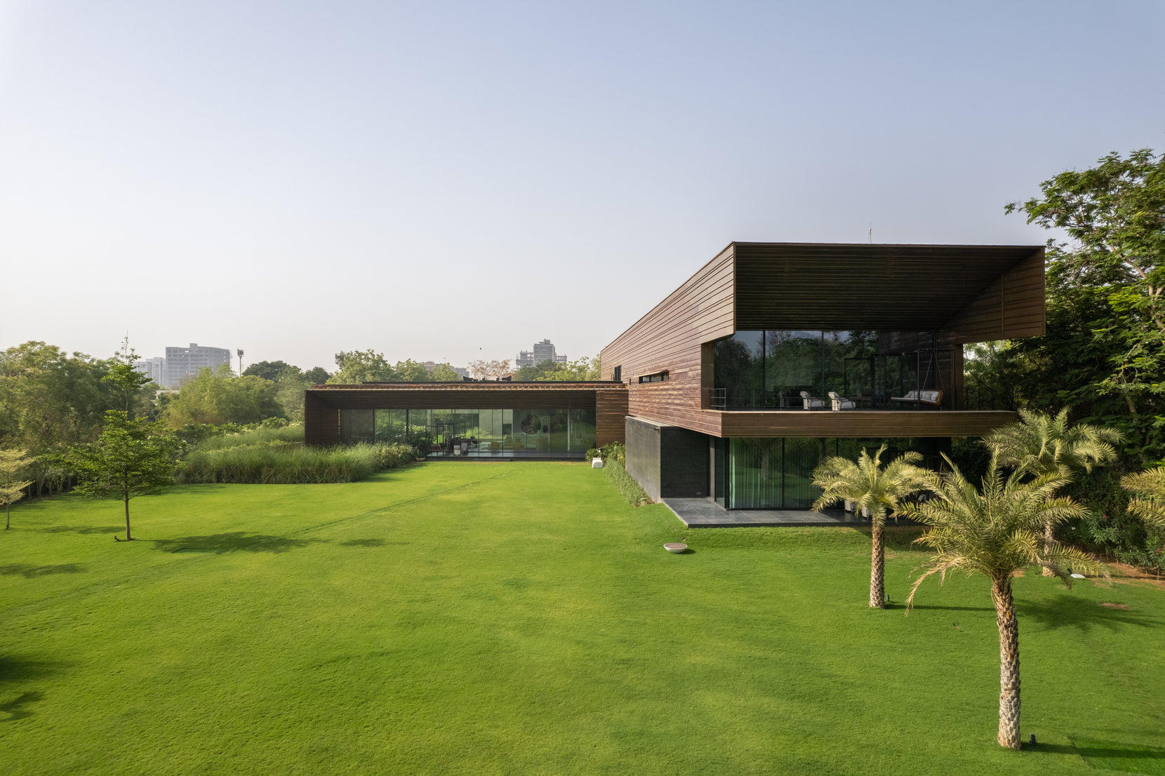 Upper timber-clad volume cantilevered over the ground floor, seen across the lawn with palms