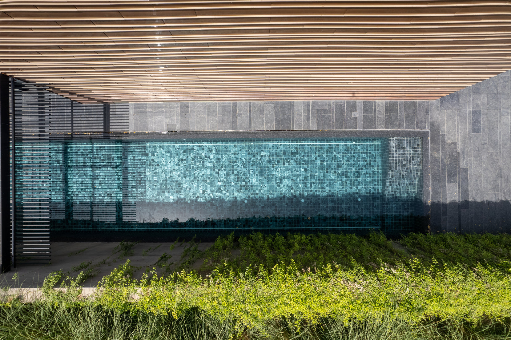 Turquoise-tiled water wall behind a louvred canopy, grasses in the foreground