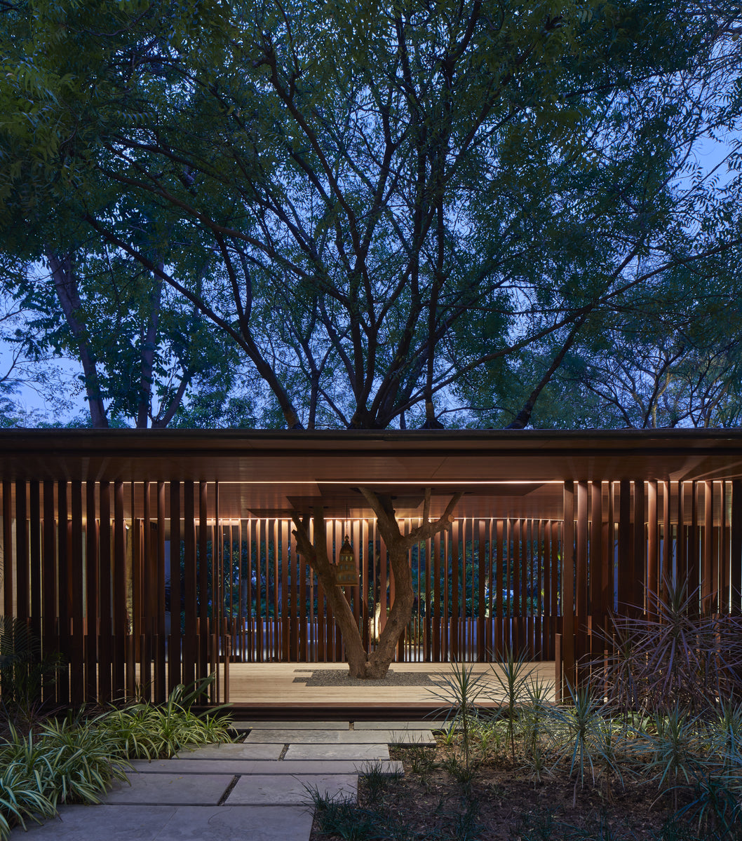 Timber-louvred pavilion lit at dusk, a retained tree rising through its deck