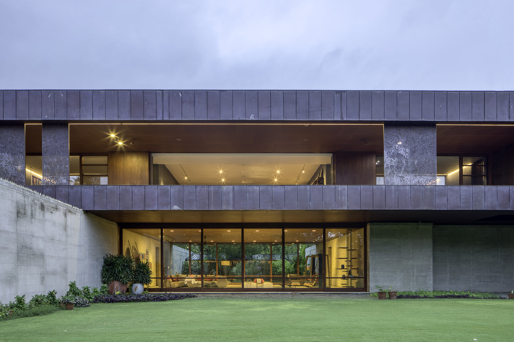 Two-storey elevation at dusk, lit interiors beneath the recessed corten upper floor