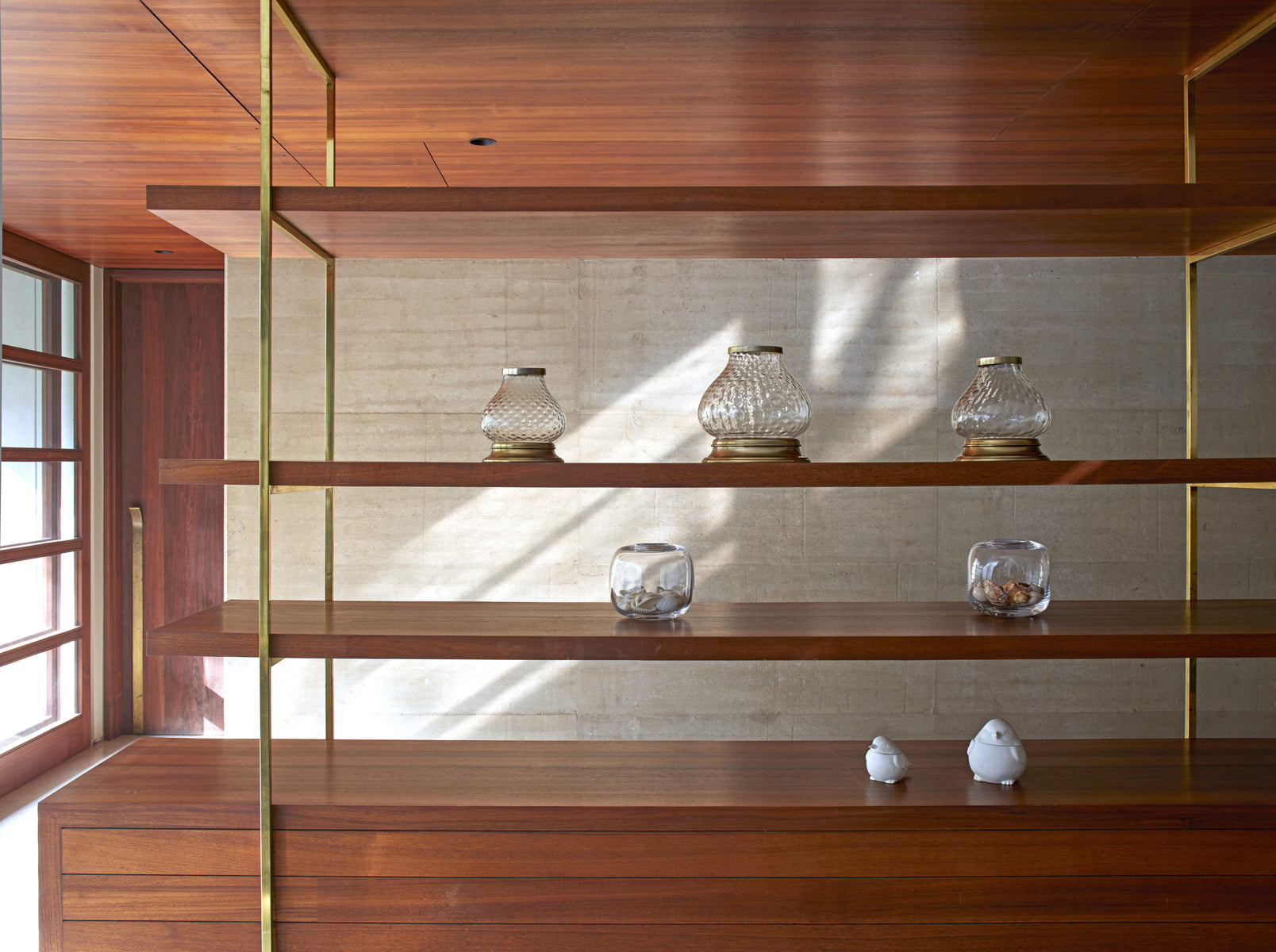 Brass-framed teak shelving holding glass vessels against a board-marked concrete wall