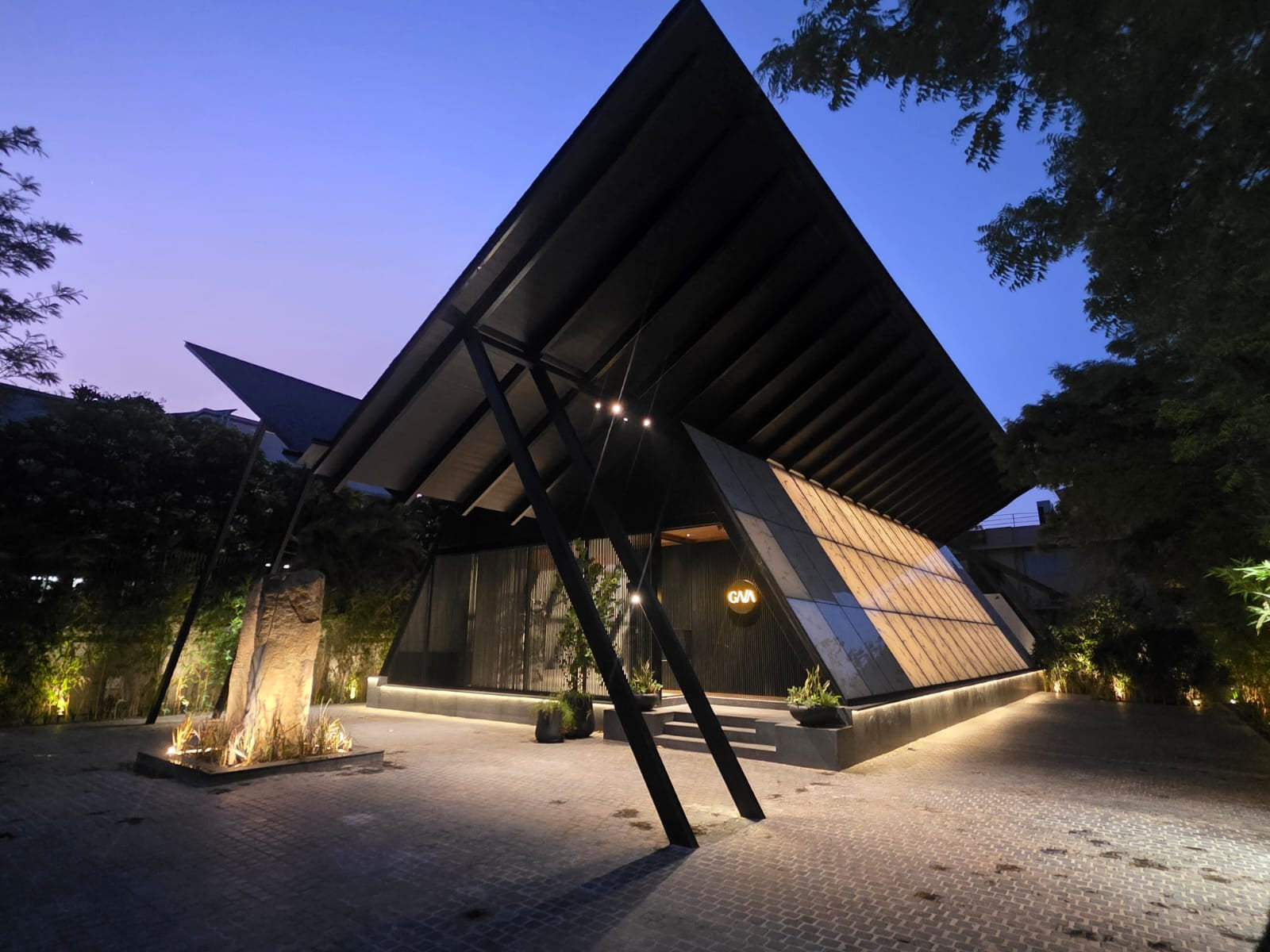Splayed steel struts carrying the winged roof at dusk, the entrance glowing beneath
