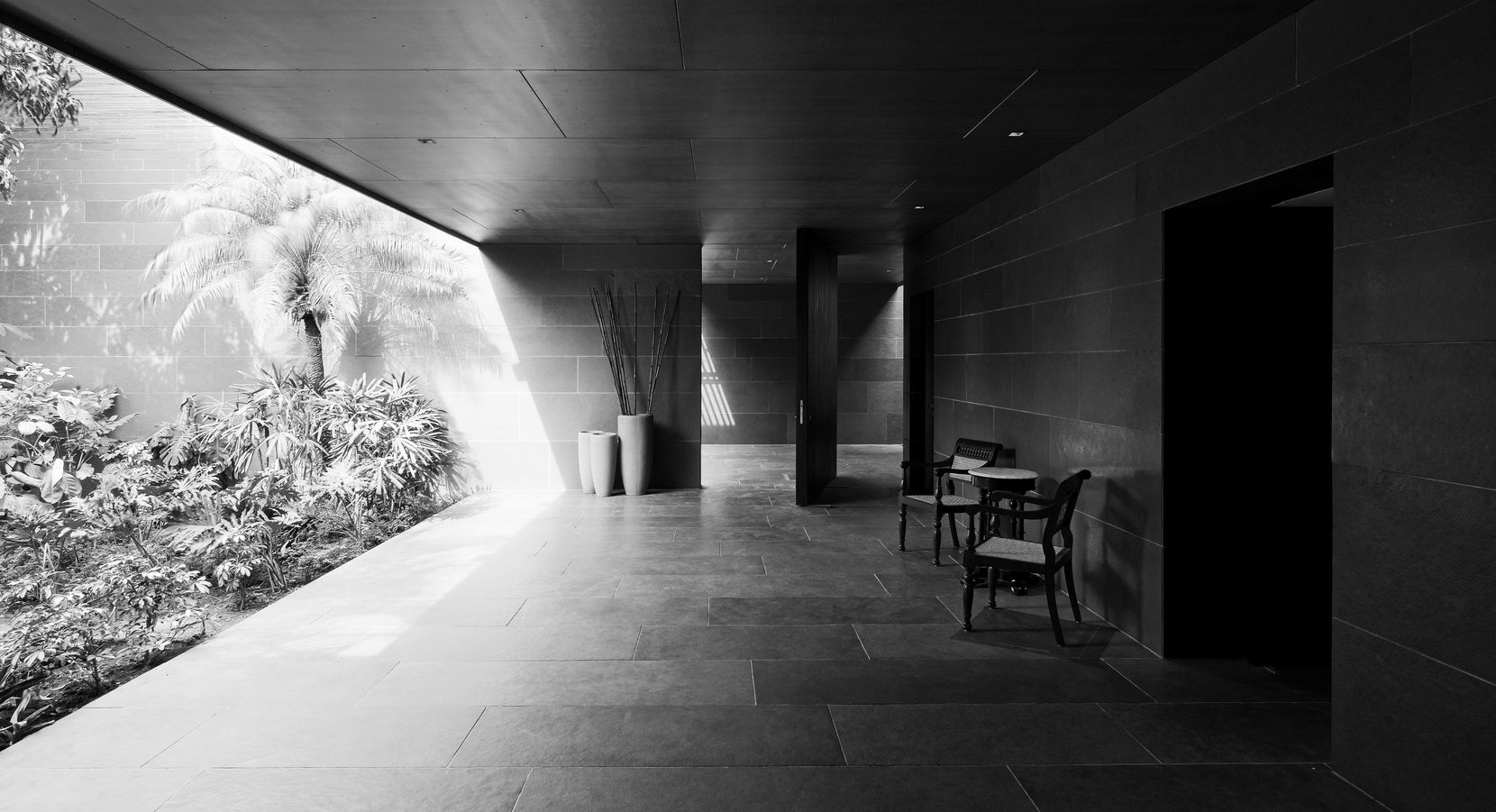 Shaded stone verandah beside a sunken planting bed, bentwood chairs at the far end, in monochrome