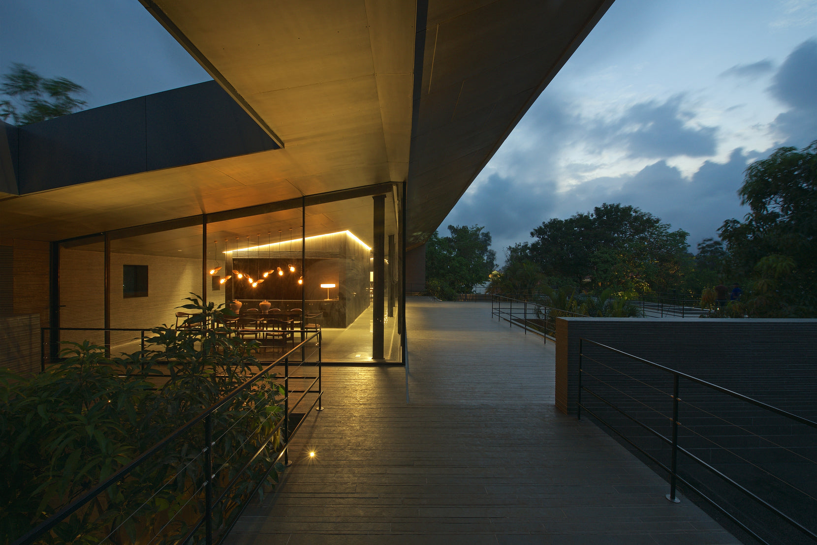 Lit dining room under a deep cantilever at dusk, the walkway running out towards the garden