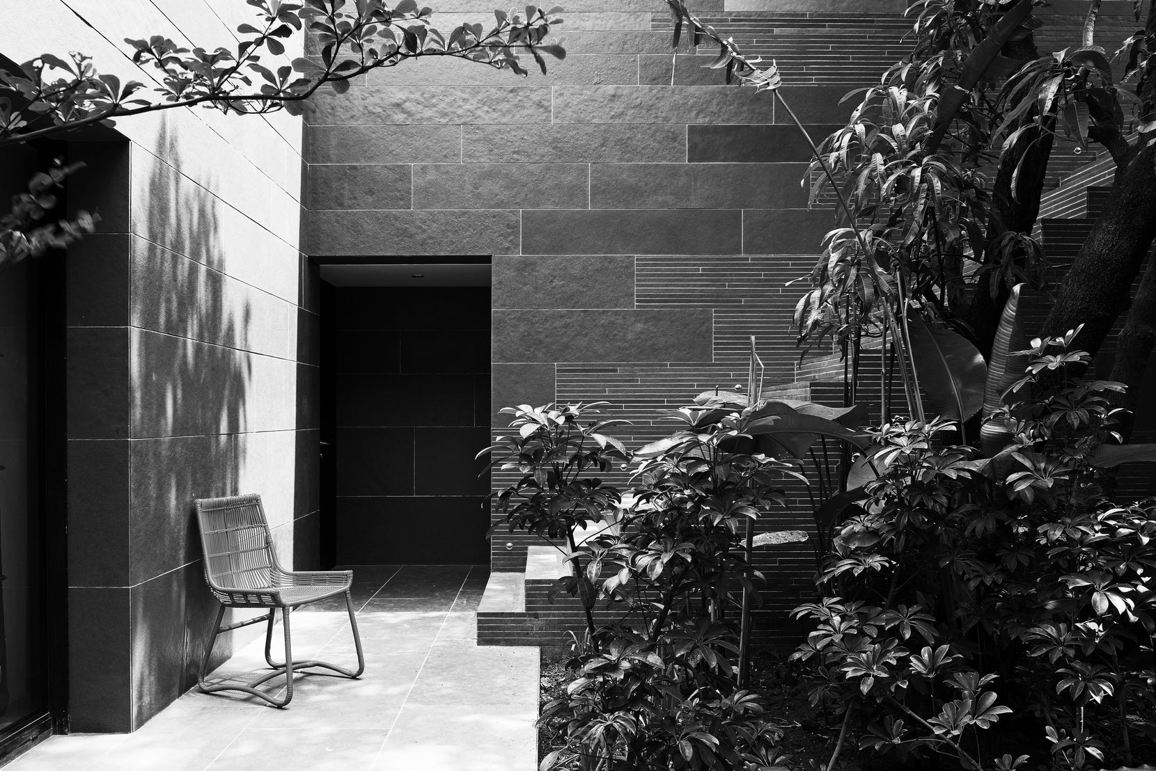 Wicker chair against a stone wall in a planted courtyard, dappled shade across the paving