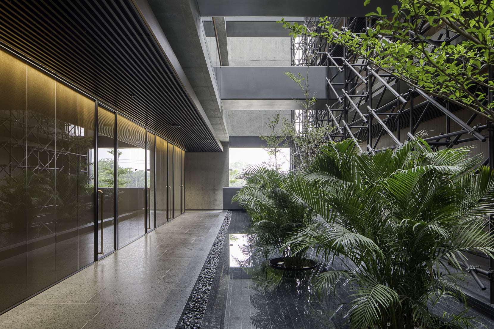 Stone corridor beside a planted water channel, tube screen rising towards the skylight