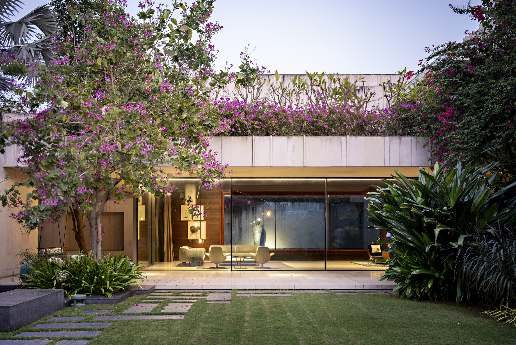 Living pavilion glowing at dusk behind a bougainvillea tree, the lawn stepping down to it