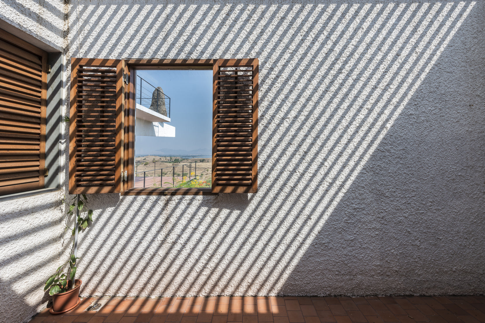 Timber-shuttered window in a rough plastered wall, striped by louvre shadows