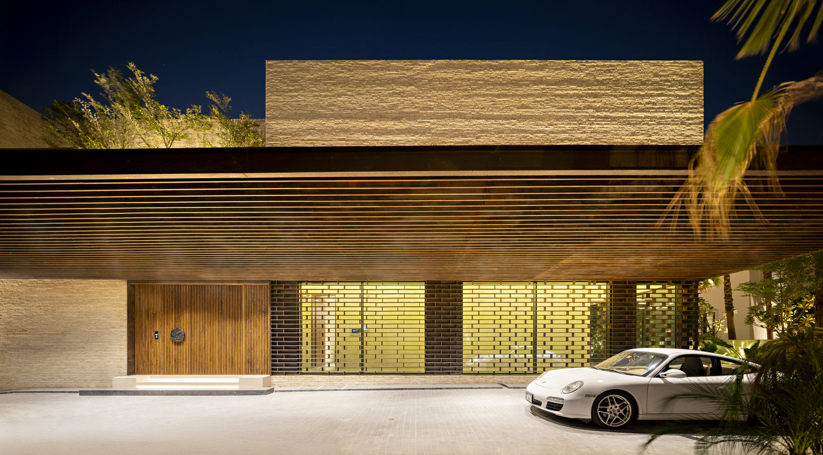 Timber louvred canopy over the entrance at night, teak door and glazed screen lit from within