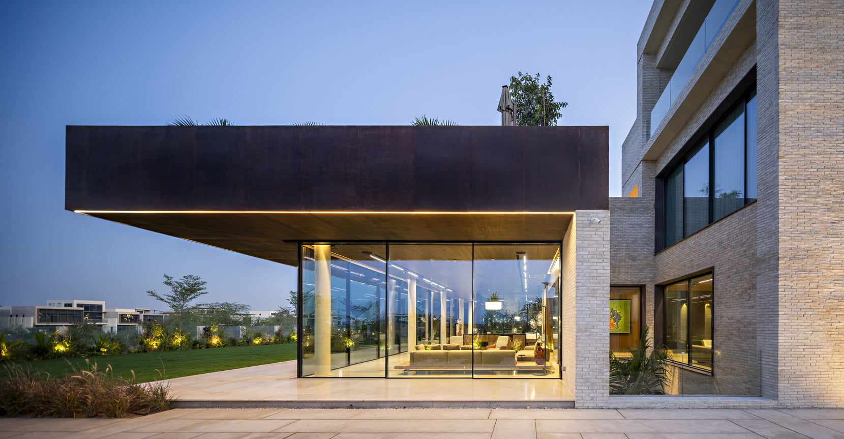 Corten-clad cantilever sheltering a glazed pavilion at dusk, lawn stretching beyond