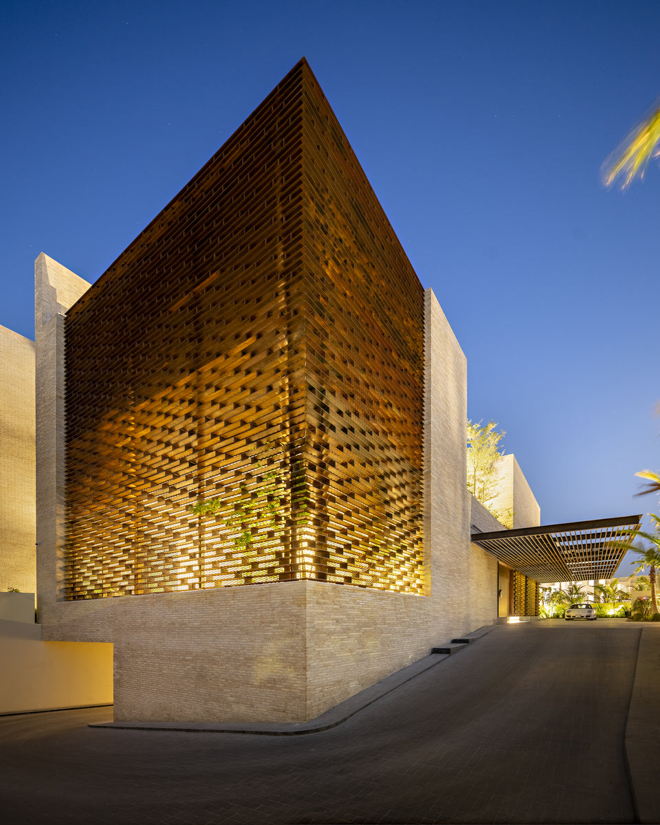 Lattice-screened corner block lit from within, driveway curving past a slatted porte-cochere