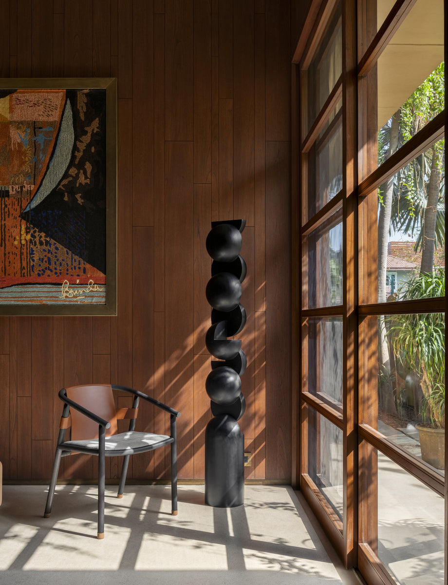 Black stacked-form sculpture and a leather chair beside a teak window, striped by afternoon shadow