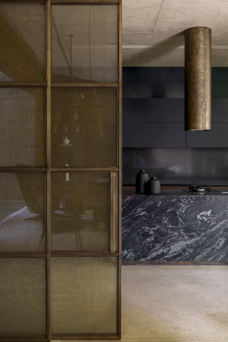 Kitchen behind a brass mesh screen, with a veined black stone island and bronze cylindrical extractor