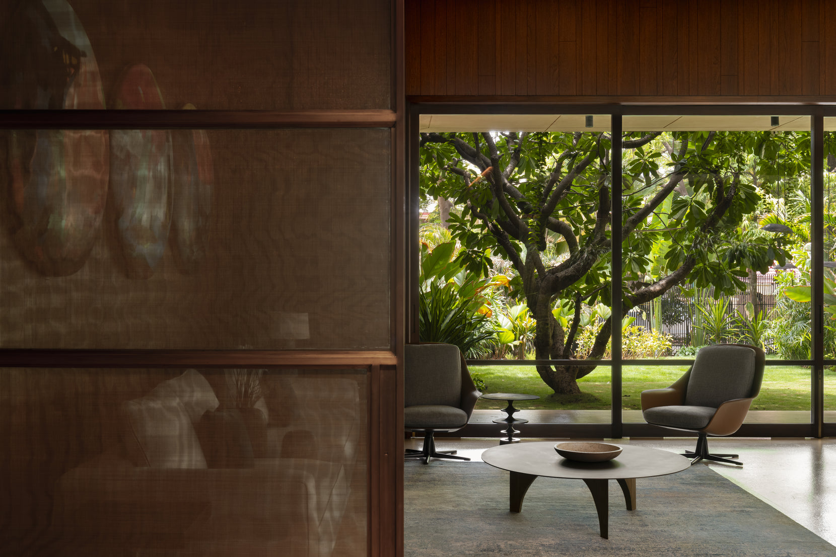 Two swivel chairs and a round table at the glass wall, a mature tree on the lawn beyond
