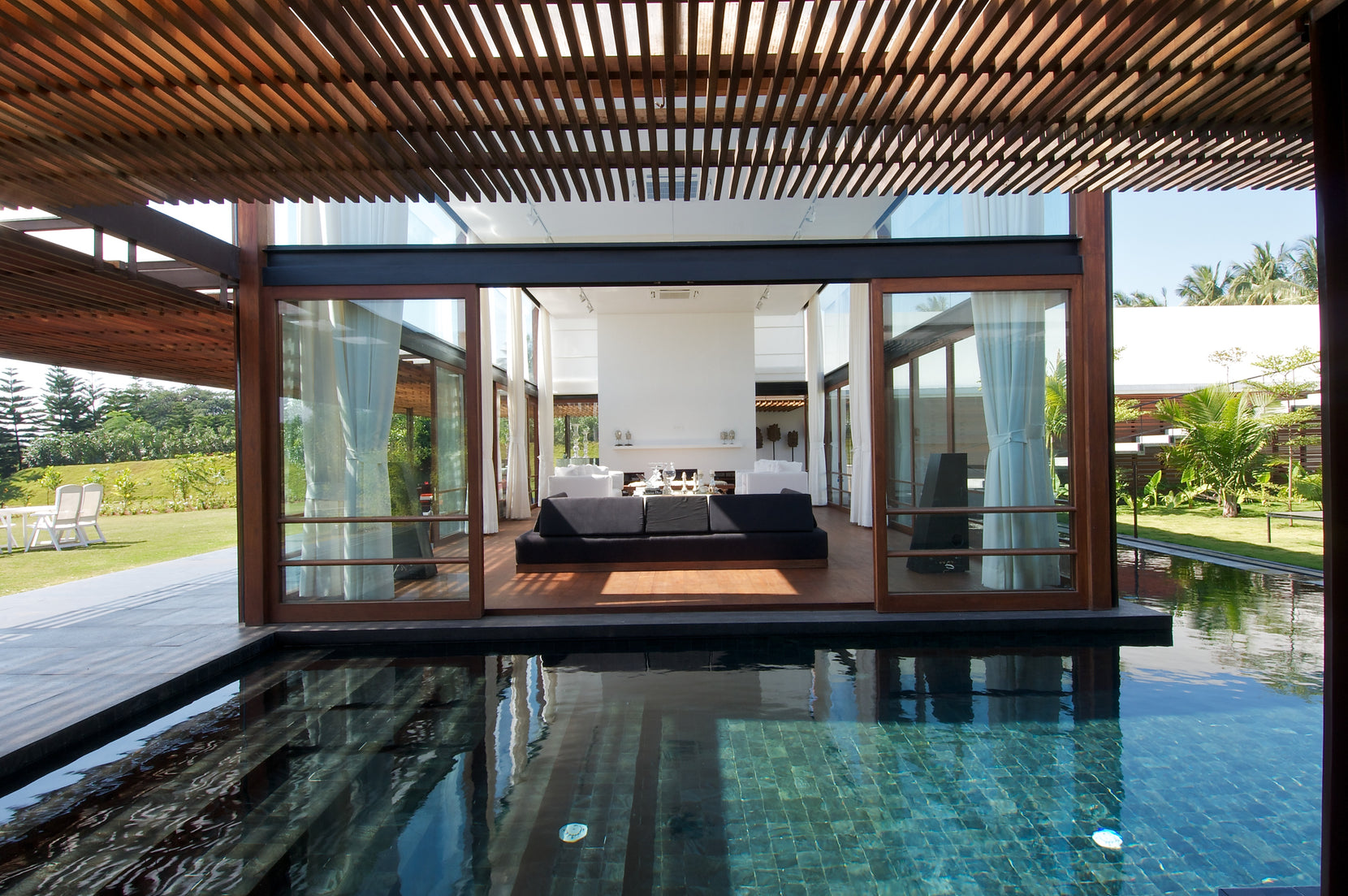 Glass living pavilion framed in teak, reflected in the still water of the pool