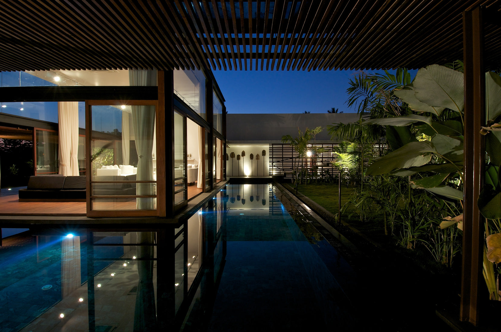 Pool at night between the lit glass room and dense planting, underwater lights glowing