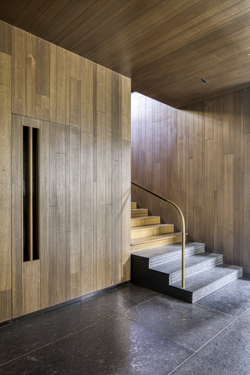 Timber-lined stair hall with a brass handrail rising over granite and stone treads