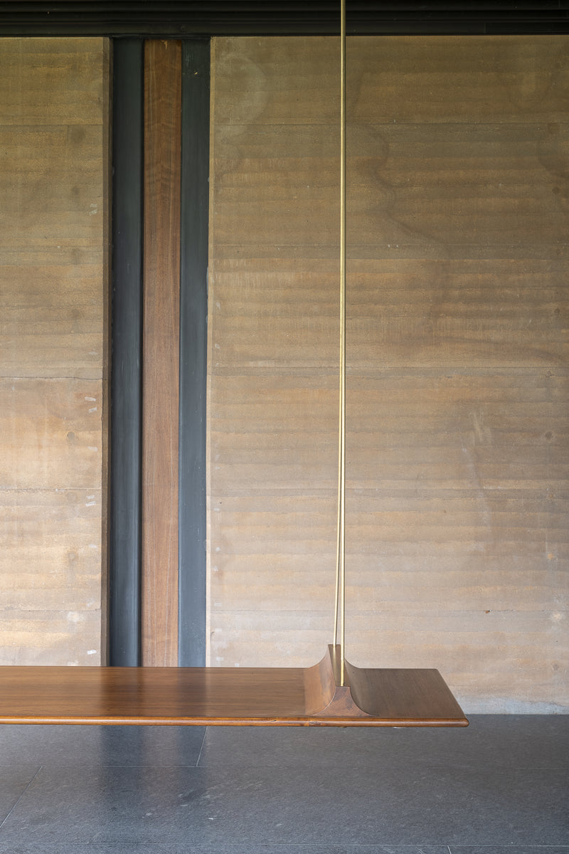 Teak swing seat suspended on a slim brass rod in front of a board-marked rammed earth wall