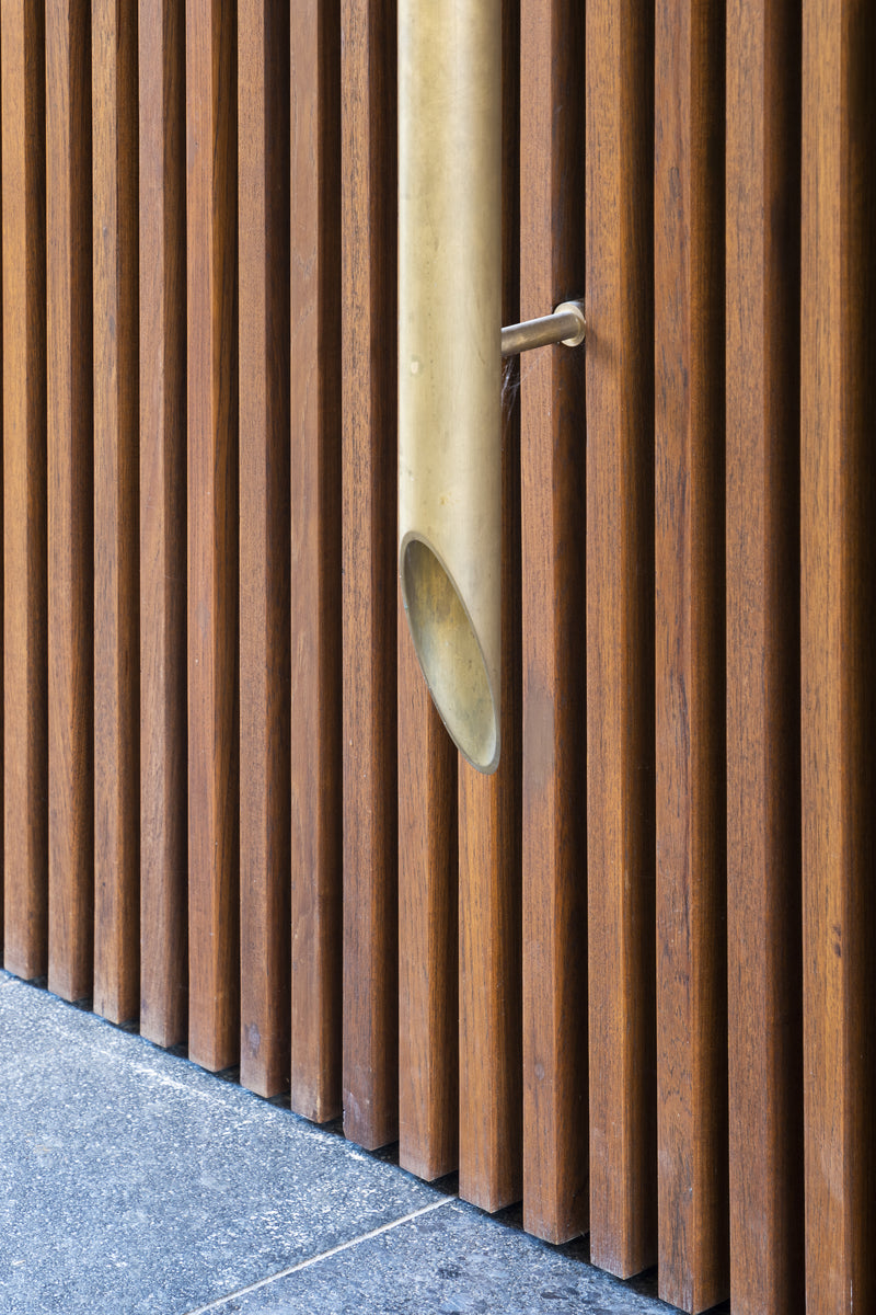 Brass rain spout emerging through vertical teak louvres above a granite plinth
