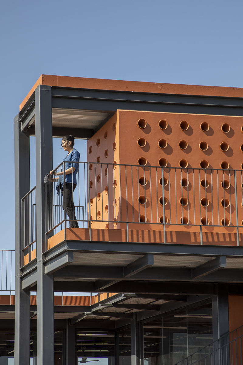 Student on a steel balcony beside a terracotta screen of open pots against clear sky