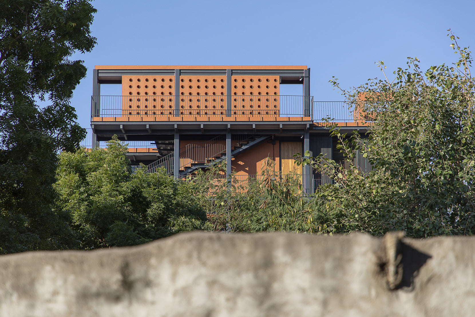 Perforated terracotta parapet and steel structure seen over a rough compound wall and trees