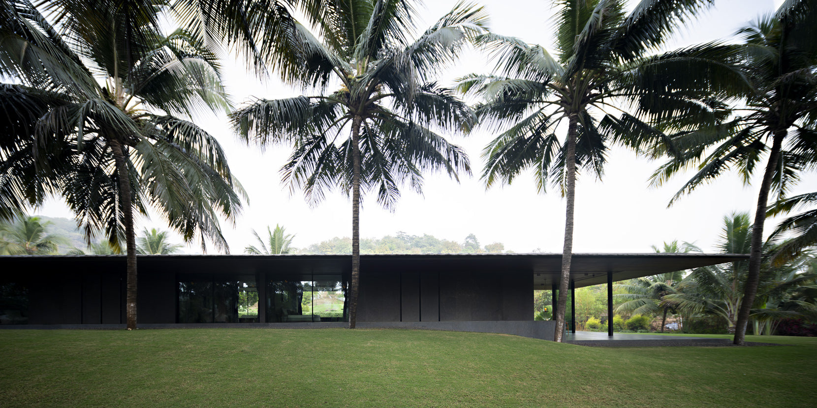 Dark stone elevation and cantilevered roof of Parikrama framed by coconut palms above the lawn