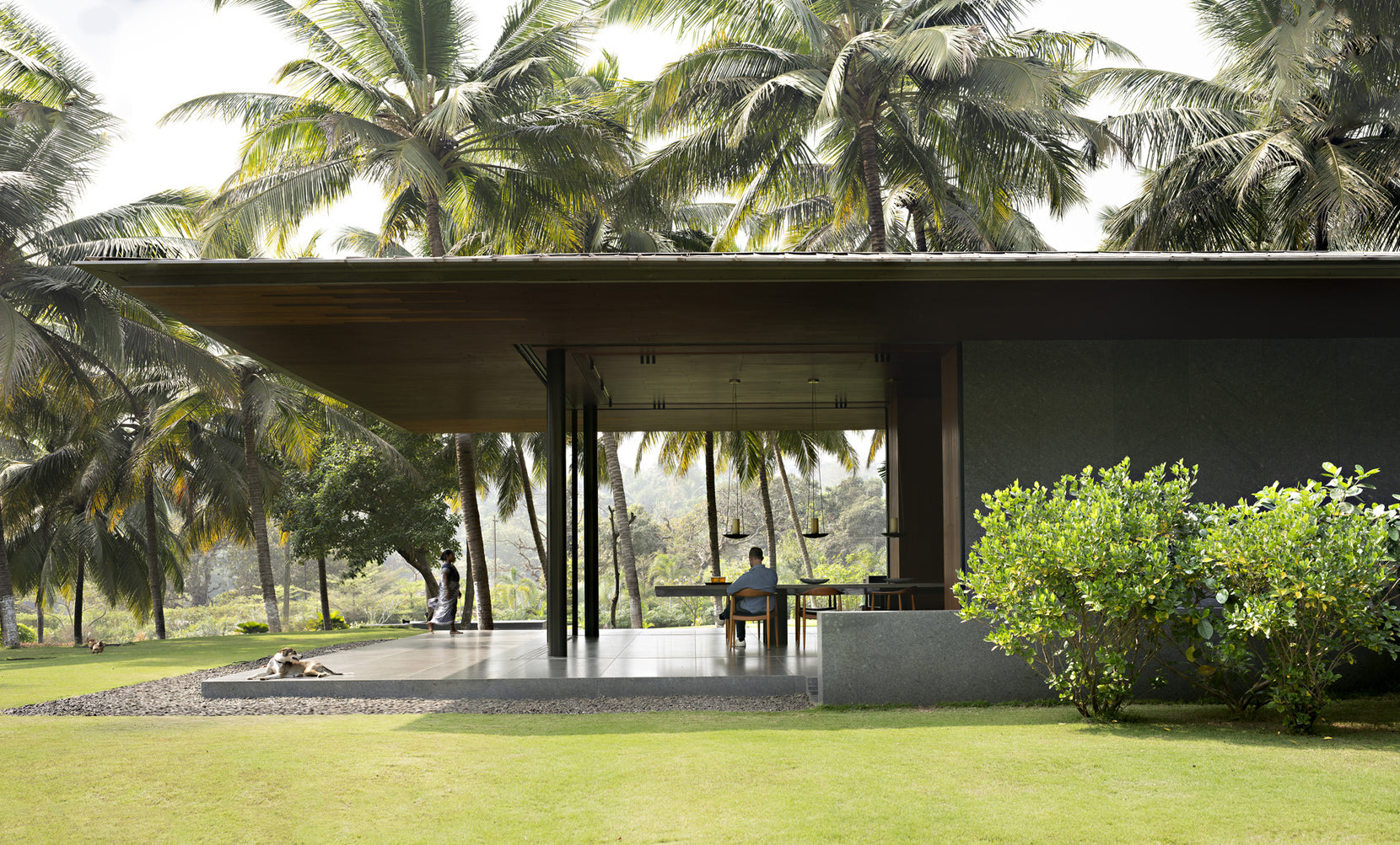 Deep cantilevered roof shading the open dining terrace, a figure seated at the table beyond the lawn