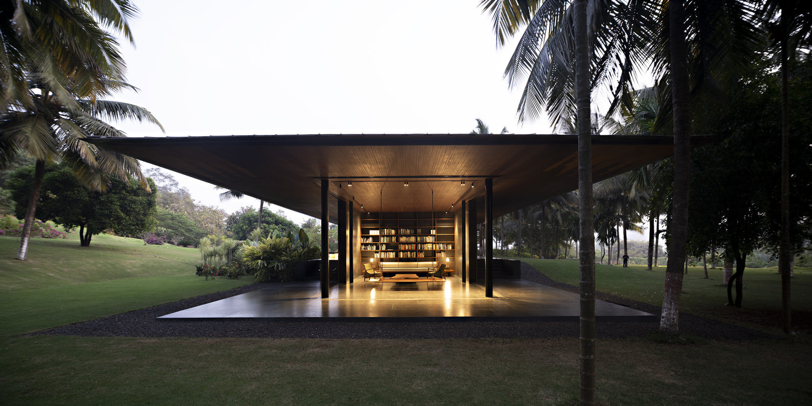 Living pavilion glowing at dusk beneath its flat roof, bookshelves lit along the far wall