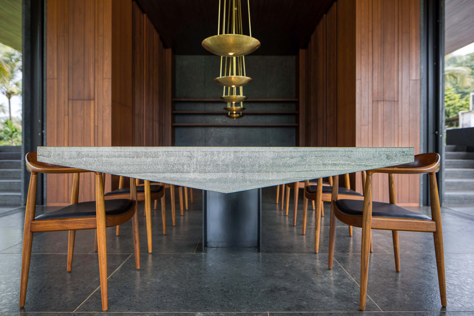 Triangular stone dining table on a single pier, teak chairs and brass pendants above
