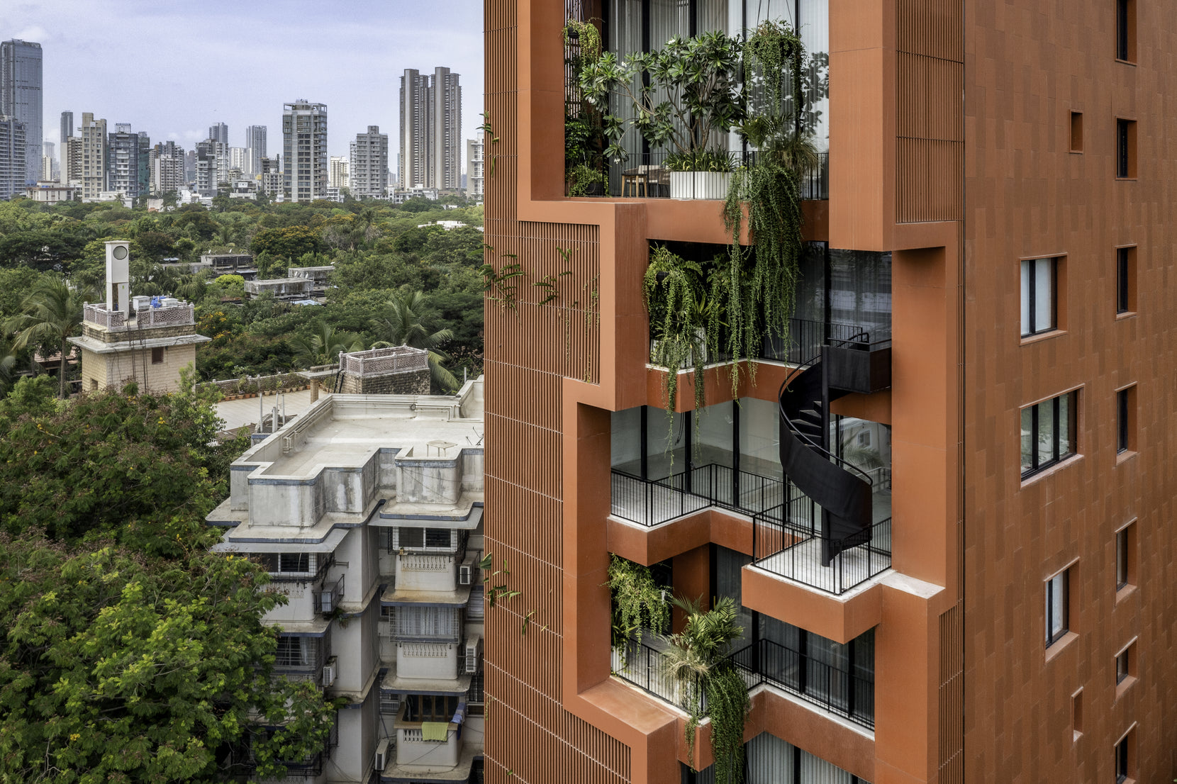 Sarvasva, a terracotta-clad tower in Mumbai, its planted balconies and black spiral stair above leafy roofs