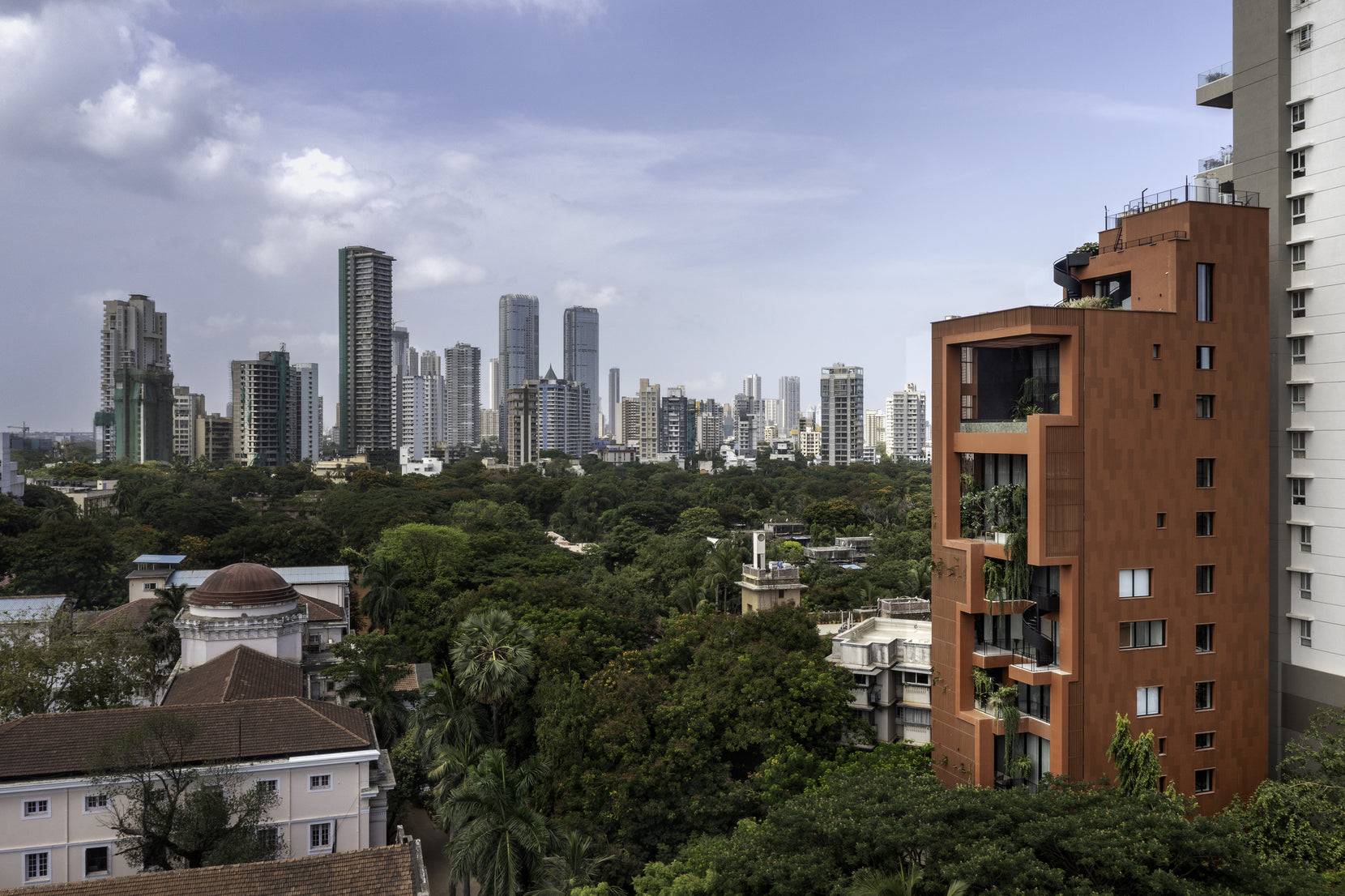 Terracotta tower at the edge of a green Mumbai neighbourhood, the high-rise skyline stretching behind
