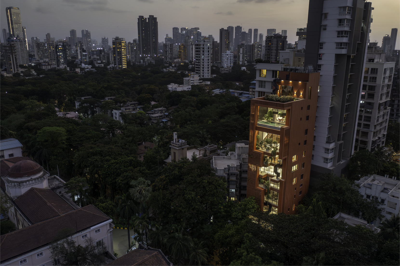 Sarvasva lit from within at dusk, stacked terraces glowing above a dark tree canopy