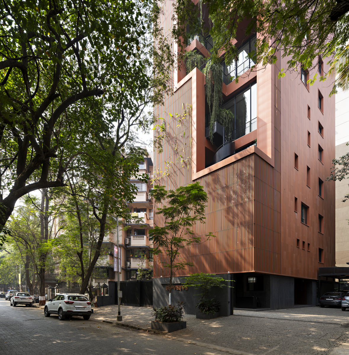 Street corner at Sarvasva, fluted terracotta screens over a dark stone base beneath rain trees