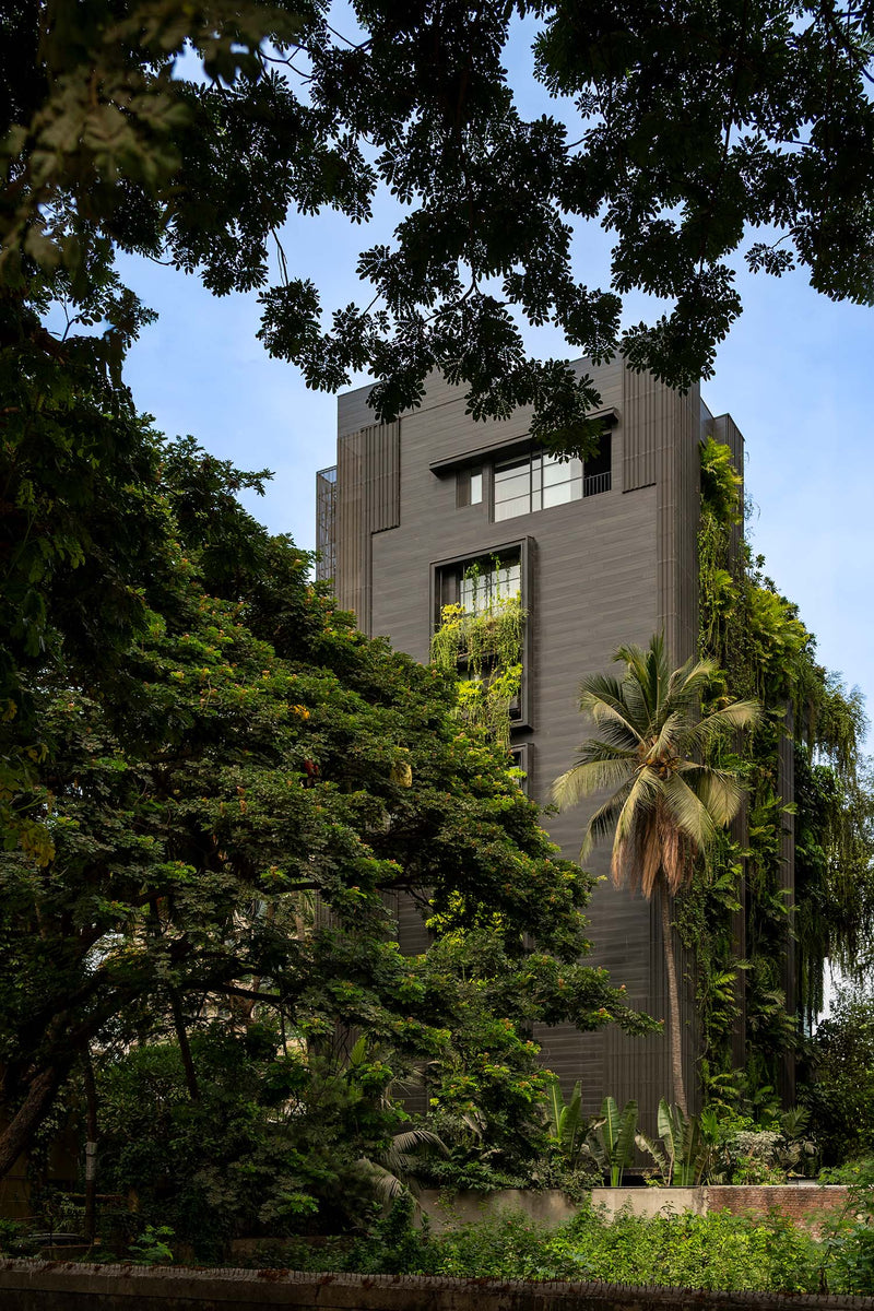 Shiv Kunj rising behind rain trees and a coconut palm, dark ribbed cladding and planted recesses