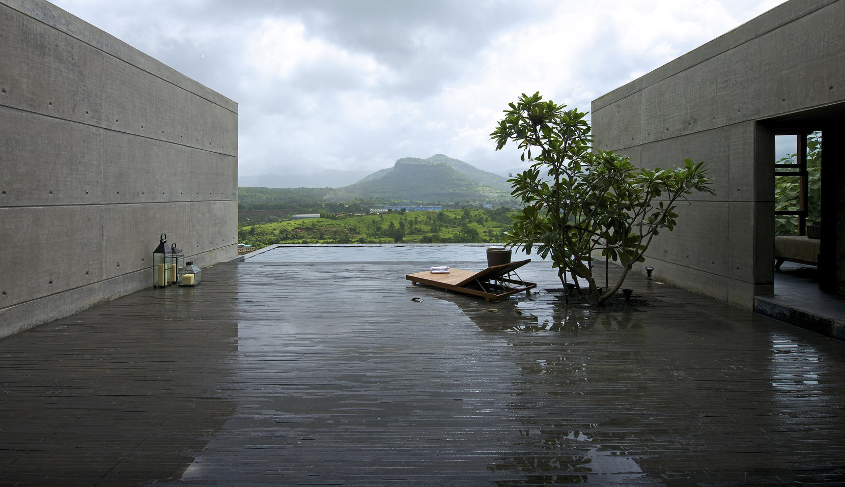 Rain-wet stone deck and infinity pool framed by two concrete walls, frangipani and a daybed